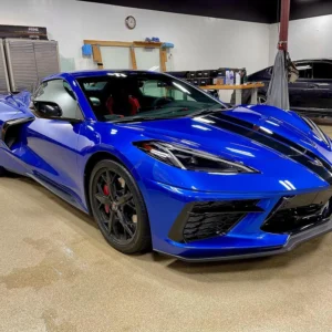 Un Chevrolet Corvette azul brillante con franjas de carreras negras, aparcado en un taller de detallado, con llantas negras, pinzas de freno rojas y un acabado brillante bajo la luz del techo.