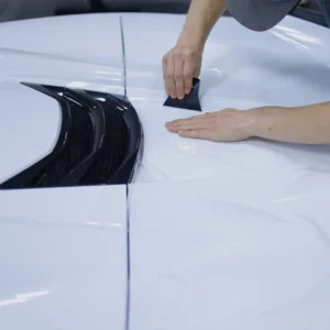 Primer plano de unas manos utilizando una espátula para aplicar una lámina protectora transparente sobre el capó de un coche blanco.