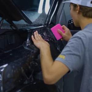 Un técnico utiliza una espátula rosa para alisar la lámina protectora de pintura sobre el guardabarros de un vehículo negro.