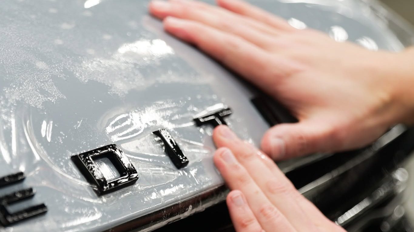 Installer smoothing XPEL paint protection film over the hood lettering of a gray SUV