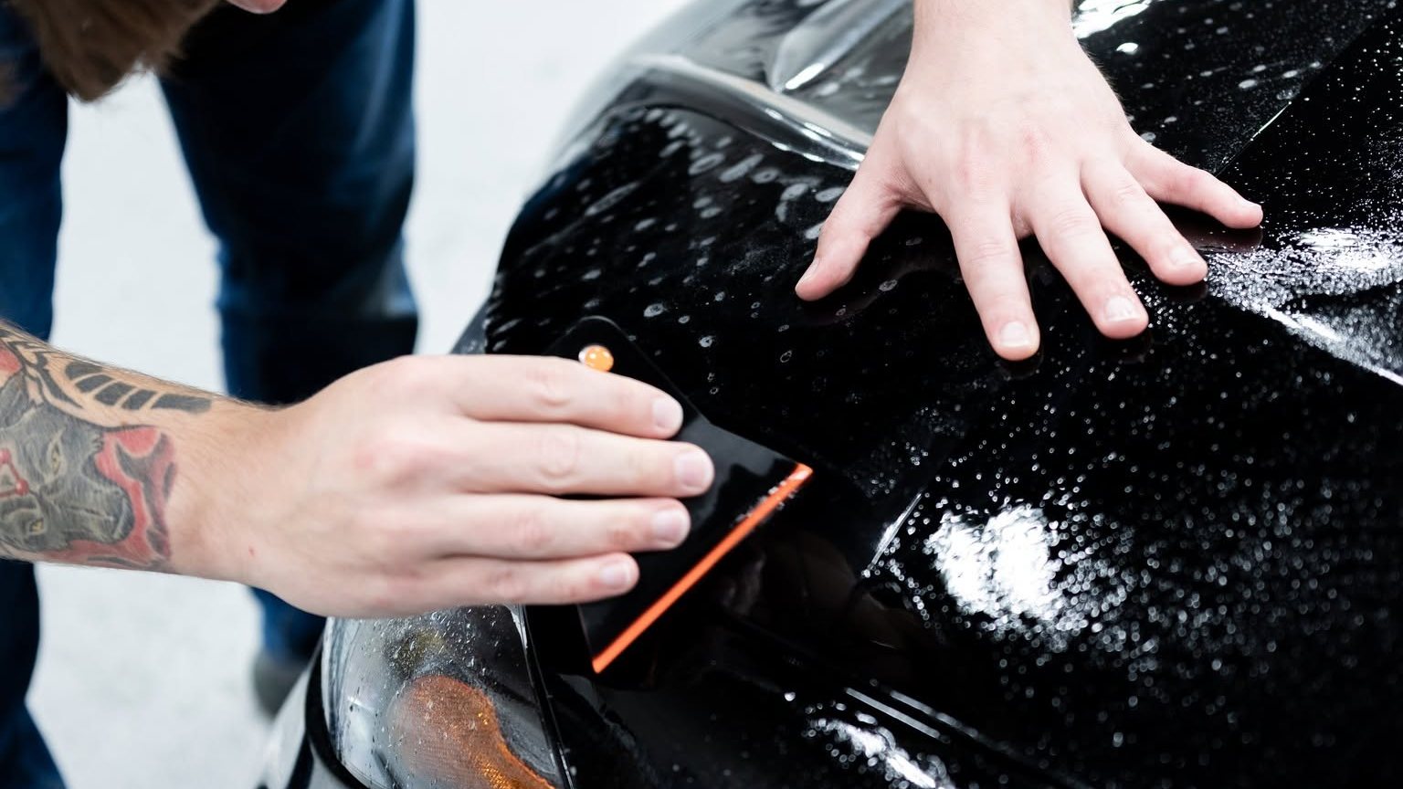 Bravo Protection installer squeegeeing XPEL paint protection film along the headlight edge of a black car